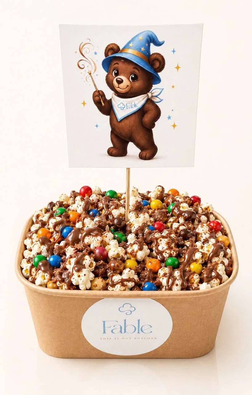 A large kraft Fable tub of Chocolate Spell popcorn with a chocolatey drizzle, dotted with round treats in bright colours, beneath an illustrated wizard bear flag wearing a Fable bandana.