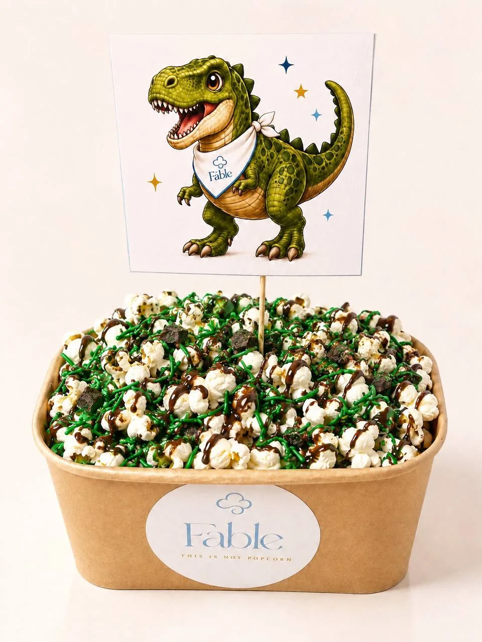 A large kraft Fable tub of Dinosaur Crunch popcorn with a brown drizzle, a dark crumble and bright green sprinkles, beneath an illustrated dinosaur flag wearing a Fable bandana.
