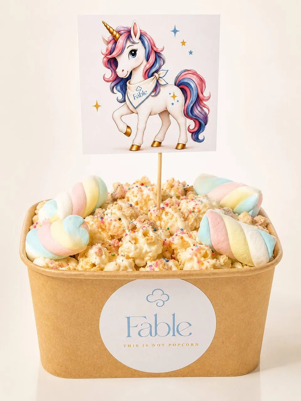 A large kraft Fable tub of Unicorn Wishes popcorn with a creamy drizzle, colourful sprinkles and pastel swirls, beneath an illustrated unicorn flag wearing a Fable bandana.